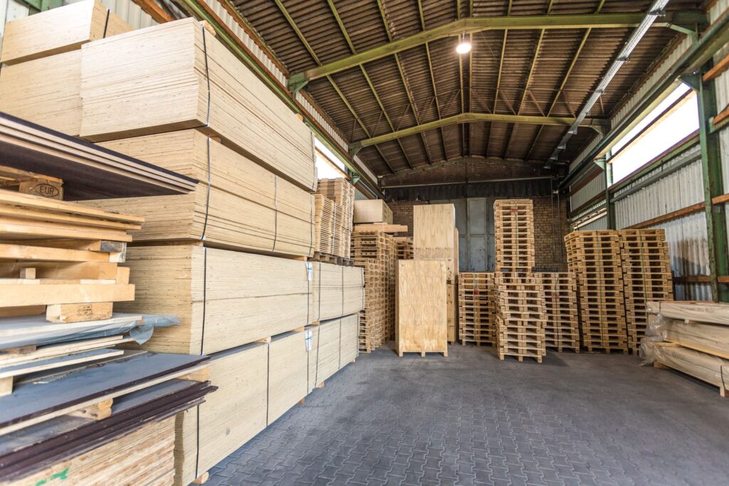 Lagerraum mit gestapelten Holzplatten und Paletten. Links hohe Stapel von Holzplatten, rechts mehrere Holzpaletten übereinander.