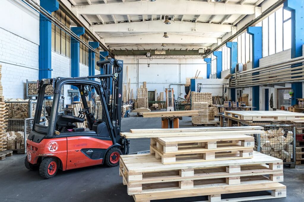 Ein roter Gabelstapler steht in einer großen Lagerhalle mit Holzpaletten. Im Hintergrund sind weitere Holzpaletten und Regale sichtbar.